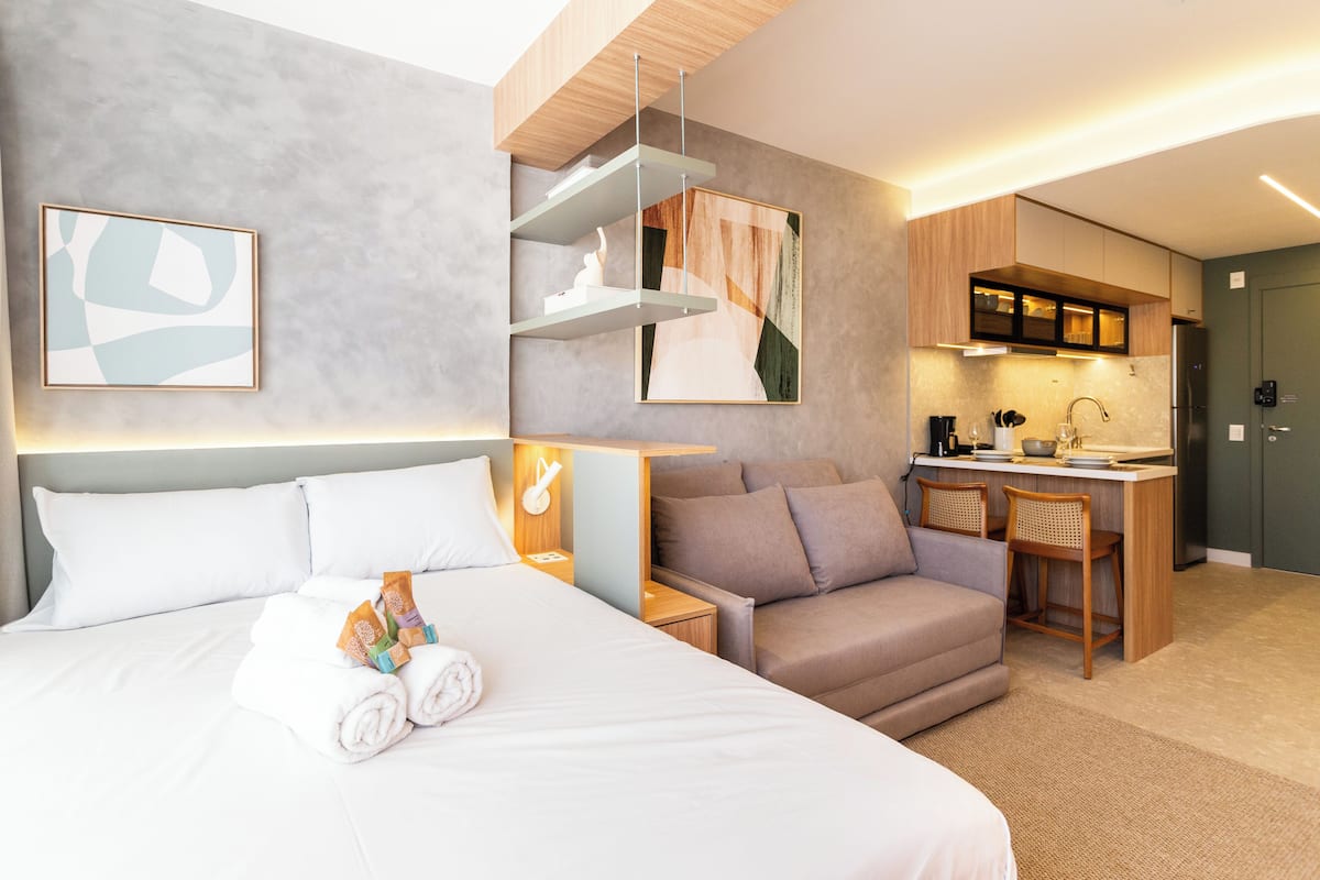 A modern hotel room with a large bed, a sofa, a small kitchen area, and a dining table.