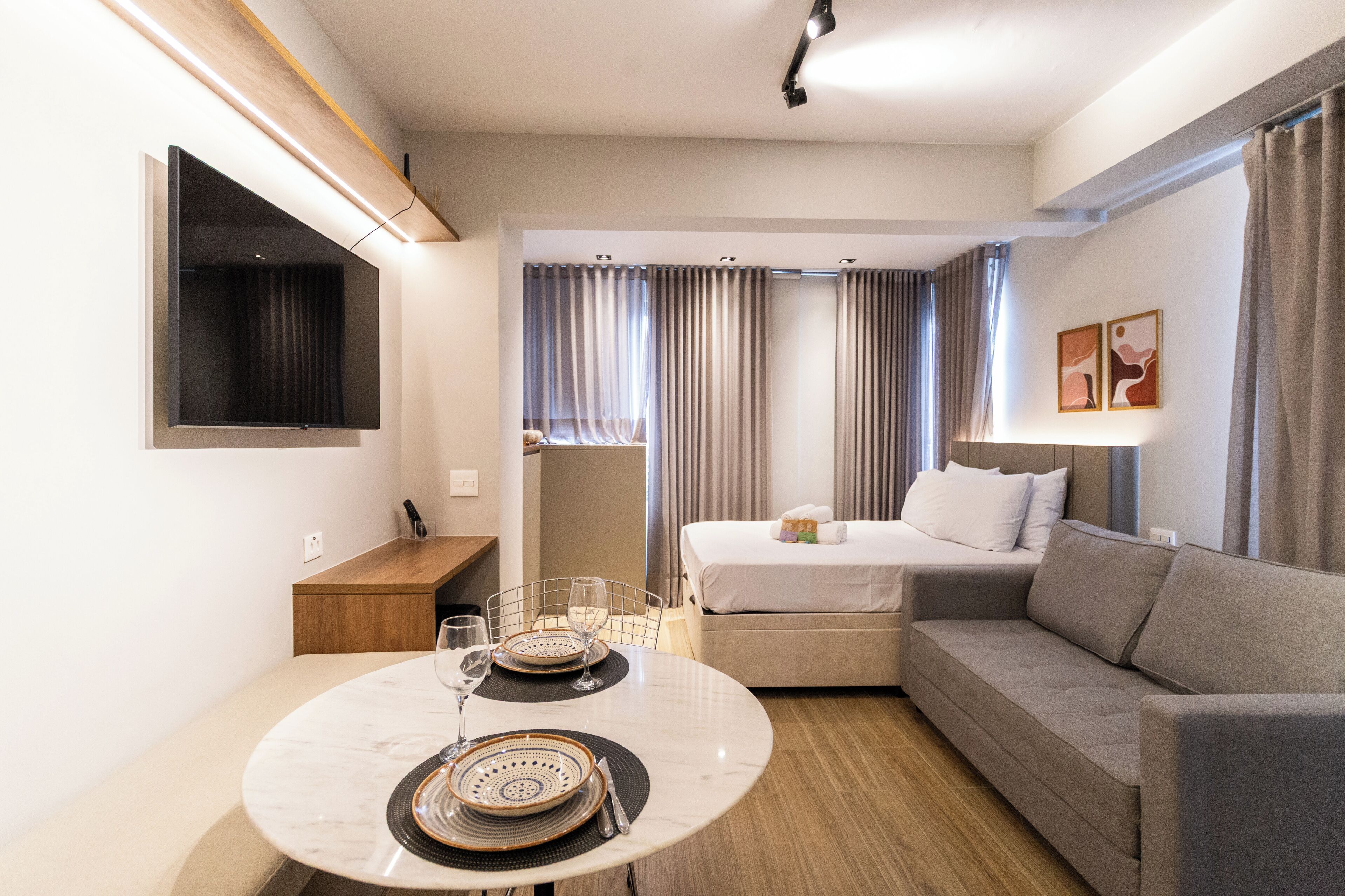 A modern hotel room with a bed, sofa, dining table, and a TV mounted on the wall.