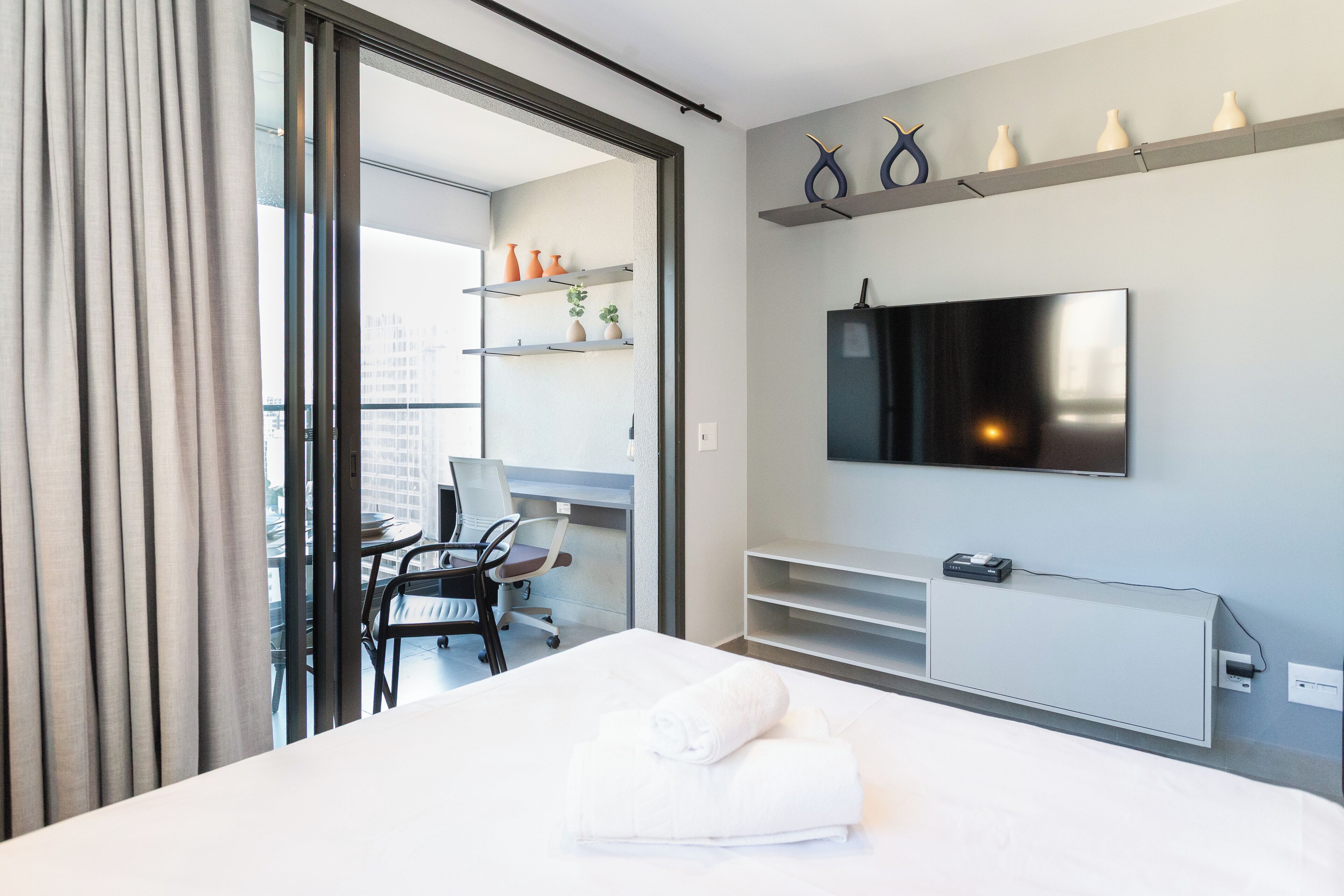 Une chambre d’hôtel moderne équipée d’un lit, d’un bureau avec un ordinateur portable, d’un téléviseur fixé au mur et d’un balcon offrant une vue sur la ville.