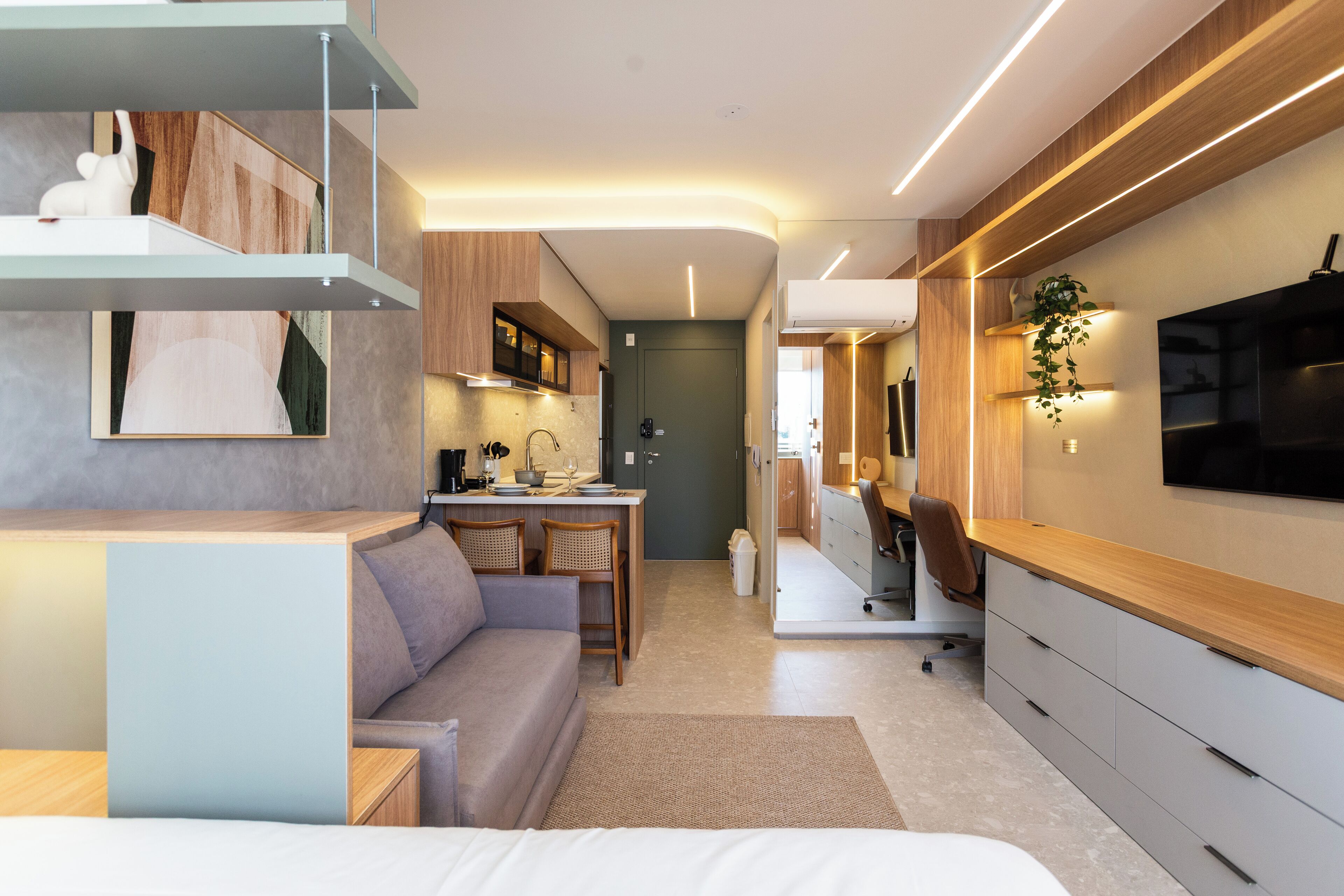 A modern, compact living space with a kitchenette, a sofa, a desk, and a TV mounted on the wall.