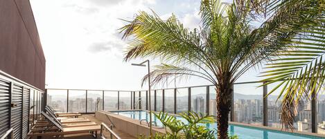 A rooftop pool area with lounge chairs, a palm tree, and a city view.