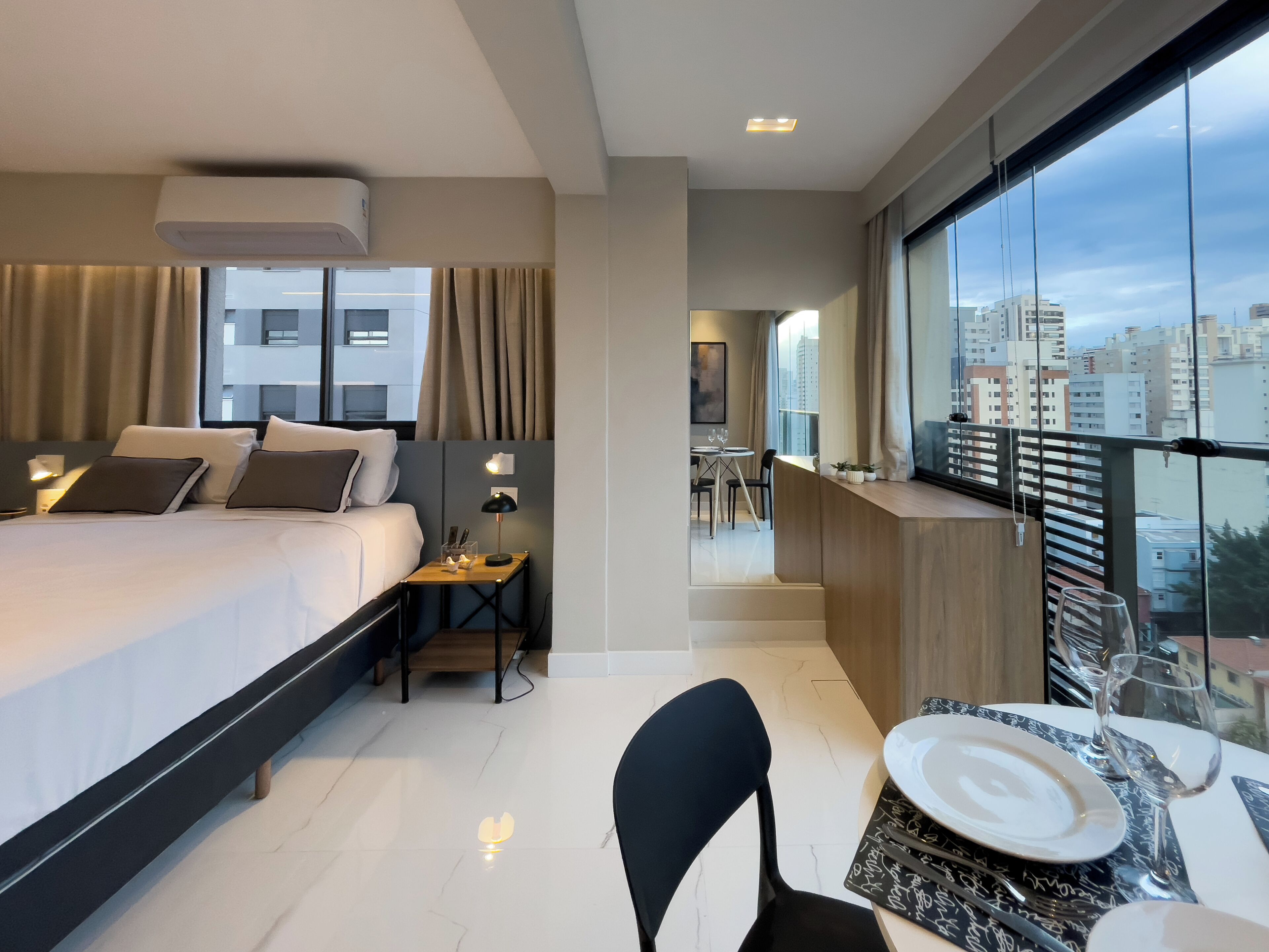 A modern hotel room with a large bed, a dining area with a table set for two, and a balcony with a city view.