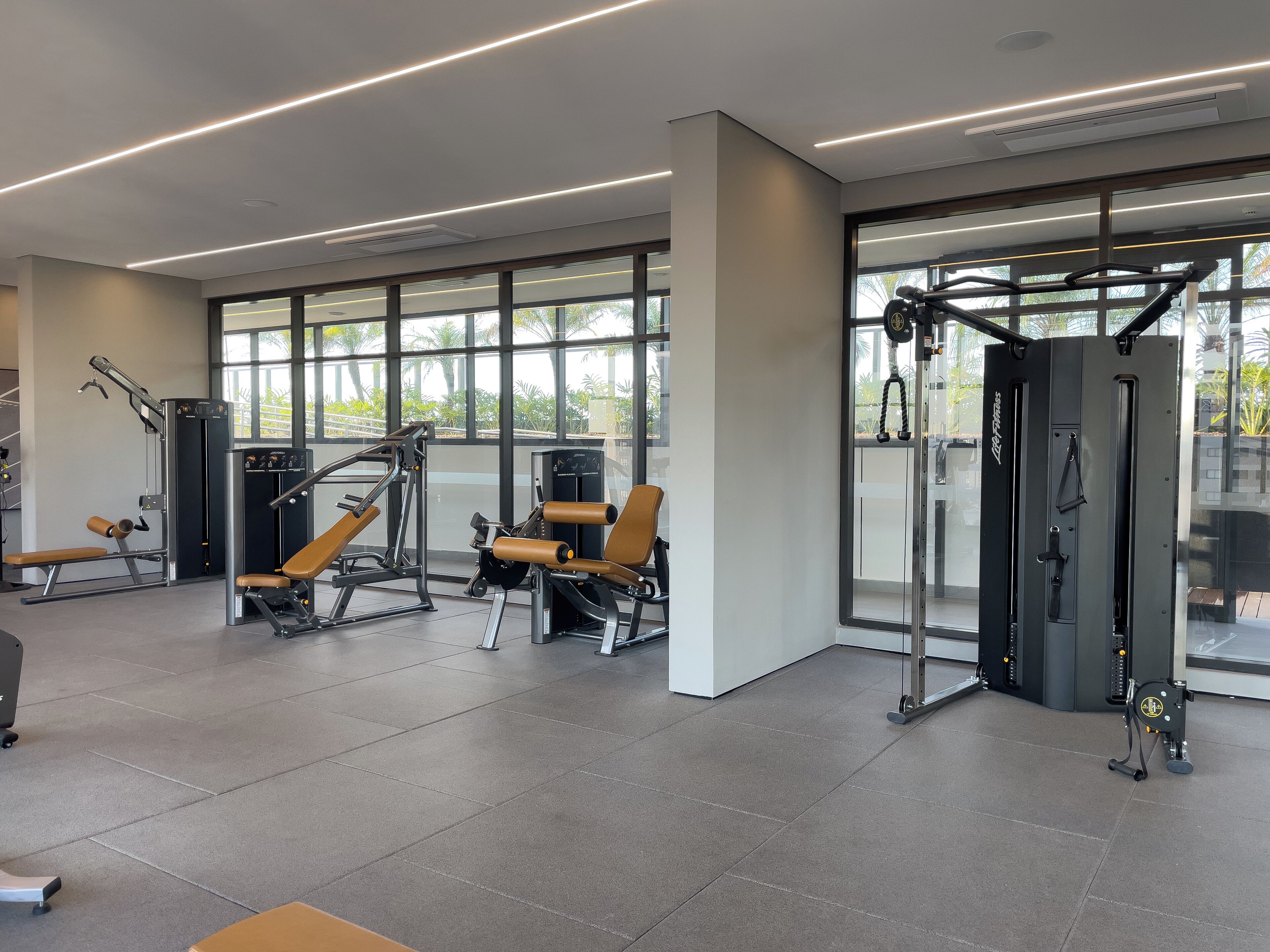 A modern gym with various exercise machines, large windows, and a clean, spacious layout.