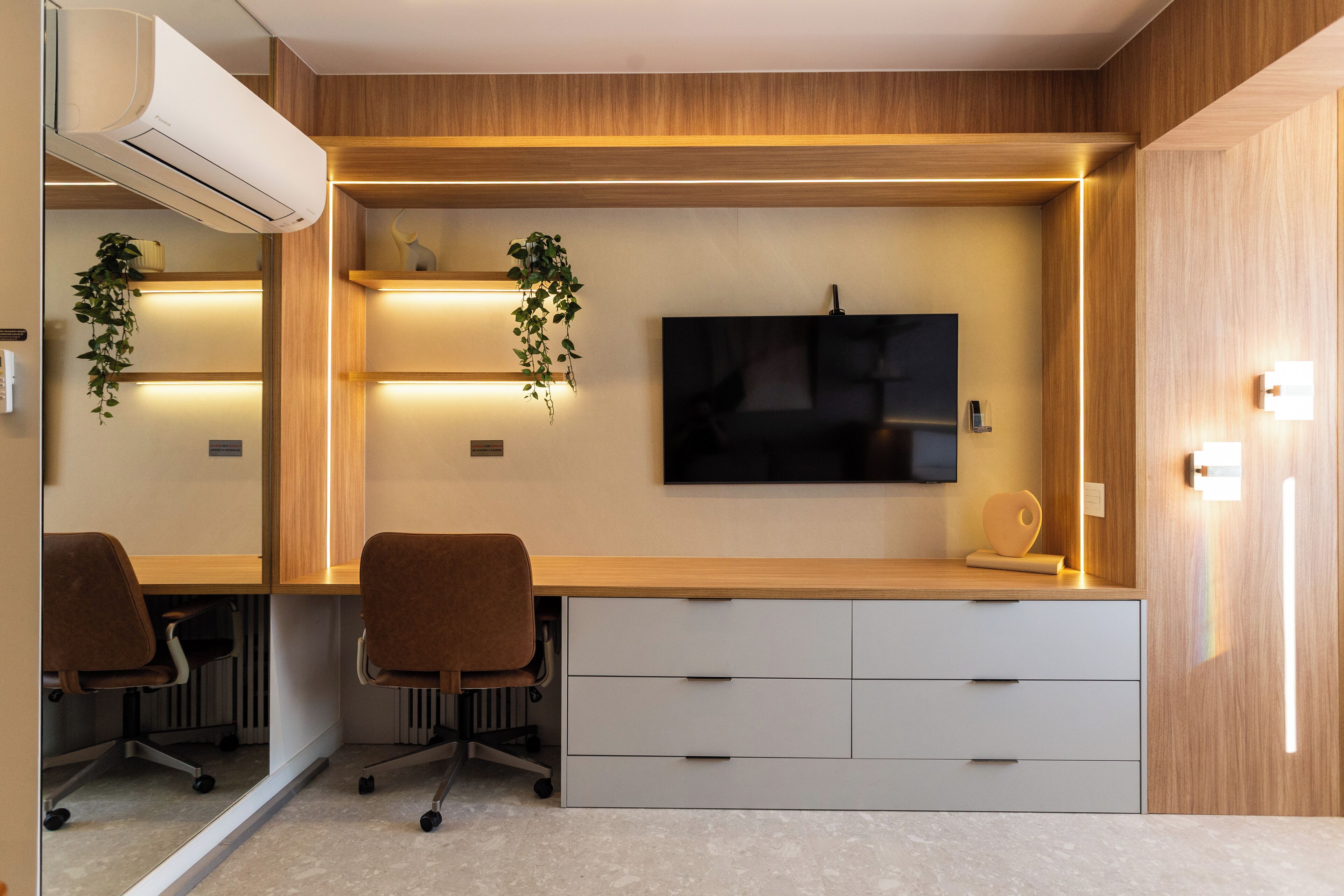 A modern office space with a wooden desk, two office chairs, a flat-screen TV, and built-in shelves with plants.