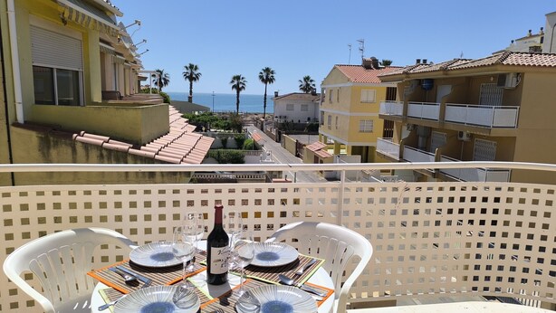 Un balcón con una mesa lista para comer, con vista a una calle costera con edificios y palmeras.