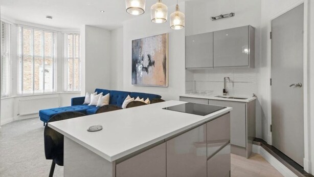 A modern kitchen with a central island, pendant lighting, and a blue sofa in a bright room with large windows.