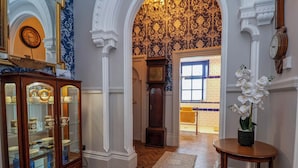 Un couloir avec une armoire en bois où sont exposées des pièces de porcelaine, une grande horloge comtoise et un papier peint à motifs floraux.