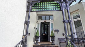 Une entrée d’immeuble avec une porte noire, une plaque de numéro et deux plantes en pot.
