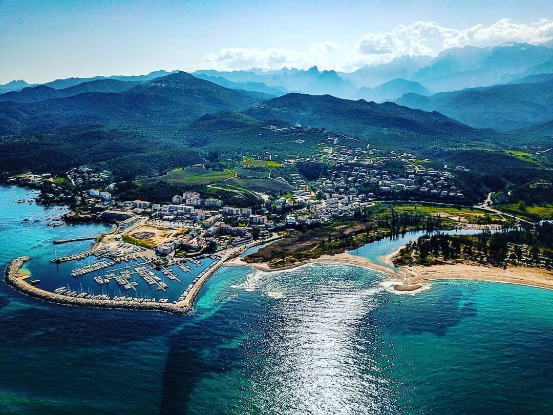 Vue aérienne d’une ville côtière avec un port de plaisance, des bâtiments et les montagnes environnantes.