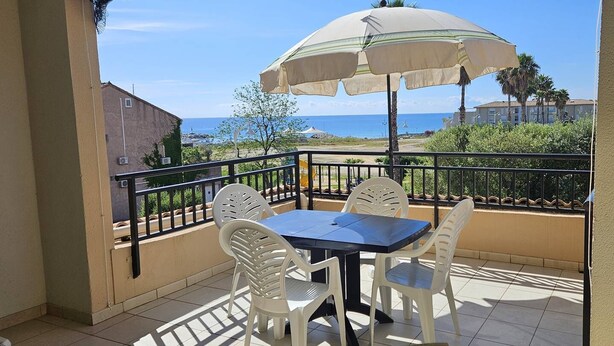 A balcony with a table and chairs, a large umbrella, and a view of the sea.