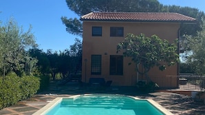 Una piscina con solárium de baldosas, un edificio con techo de tejas y vegetación circundante.