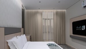 Une chambre d’hôtel moderne avec un lit soigneusement fait, un grand téléviseur à écran plat fixé au mur et une fenêtre ornée de rideaux légers.