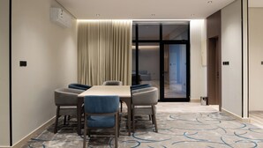 Une chambre d’hôtel moderne dotée d’un coin repas comprenant une longue table et six chaises, une grande fenêtre avec des rideaux et un tapis à motifs bleus.
