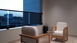 Un balcon avec deux fauteuils en osier, une petite table agrémentée d’un objet décoratif, et une vue sur les bâtiments à travers une grande fenêtre.