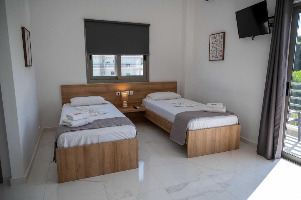 Quarto com duas camas de madeira, janela com persiana, TV de tela plana e vista para o verde lá fora.
