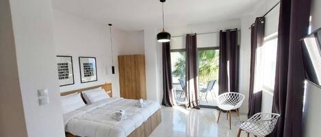 Un dormitori modern amb llit, un amb capçalera de fusta, una cadira blanca i un balcó amb cortines.
