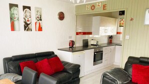 Un espace de vie compact comprenant un coin cuisine, deux canapés en cuir noir avec des coussins rouges, une table à manger en bois et une horloge murale.