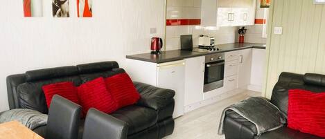 Un espacio habitable compacto con kitchenette, dos sofás de cuero negro con cojines rojos, una mesa de comedor de madera y un reloj montado en la pared.