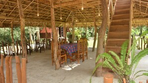 Una estructura de madera al aire libre con una mesa y sillas, rodeada de árboles y plantas.