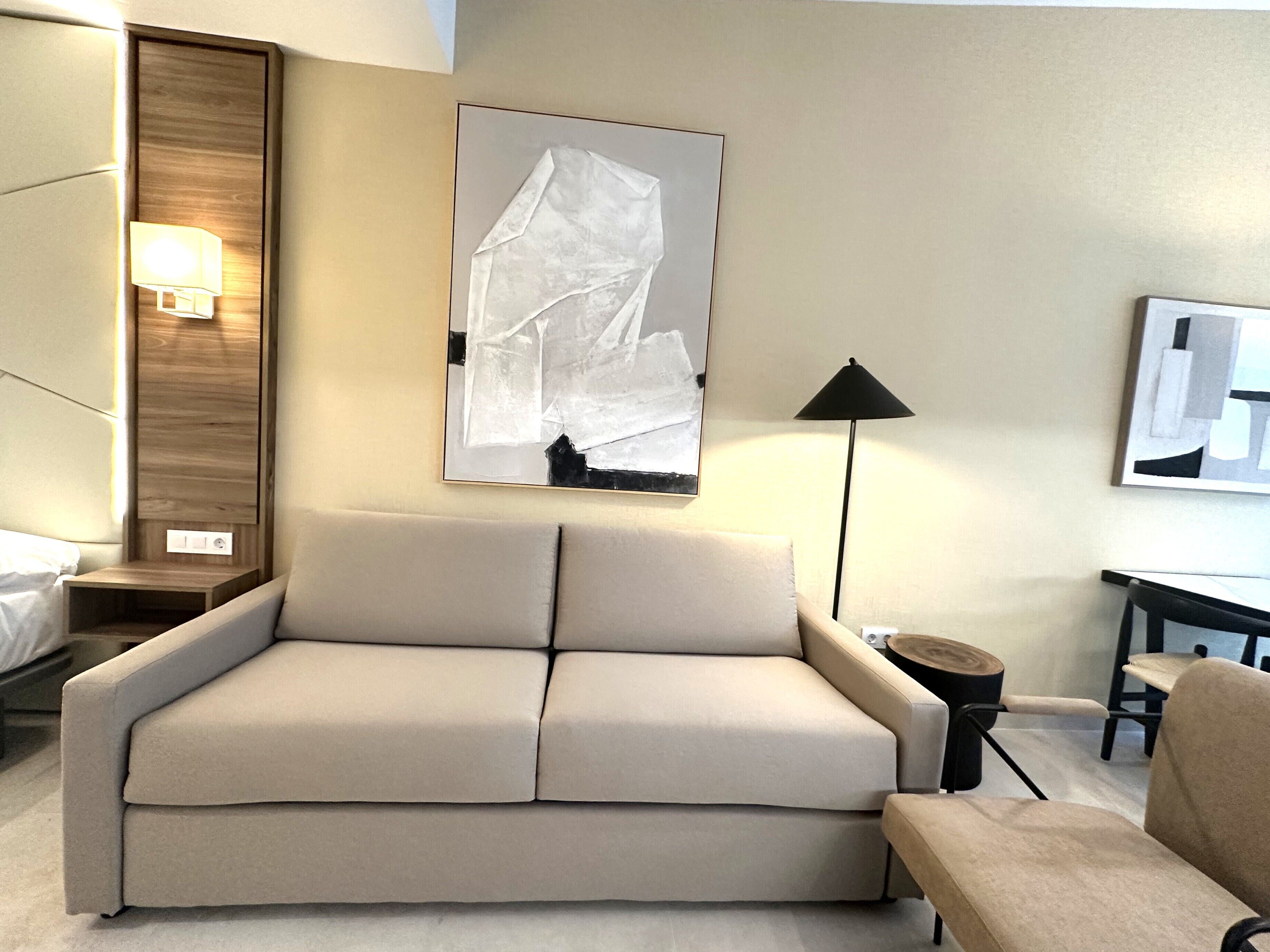 Ein modernes Hotelzimmer mit einem beigefarbenen Sofa, einem kleinen runden Tisch, einer Lampe und abstrakter Kunst an den Wänden.