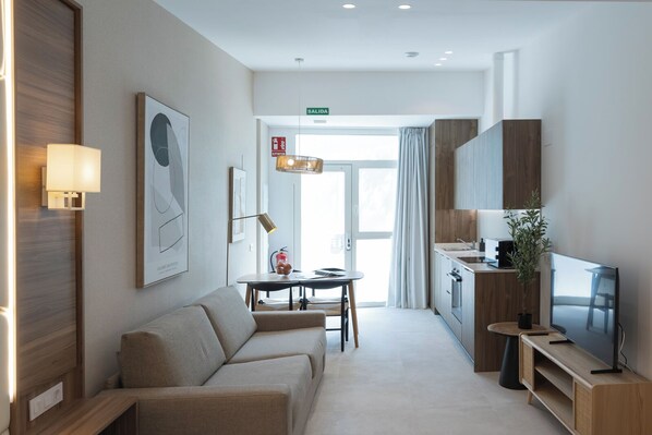 A modern hotel room with a sofa, a dining table, a TV, and a kitchen area. - MARINA AZURA (Valencia)
