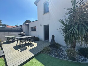 Eine Terrasse mit Tisch und Stühlen, ein Grill, ein kleiner Garten mit einer Palme und ein weißes Gebäude mit Tür und Fenster.