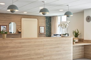 A reception area with a wooden reception desk, pendant lighting, framed pictures, and potted plants. - Quality Aparthotel Saumur (Saumur)
