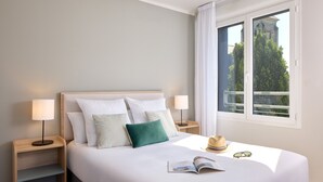 A neatly made bed with white linens and multiple pillows, a bedside table with a lamp, a window with a view of greenery and a building, and a hat and sunglasses on the bed. - Quality Aparthotel Saumur (Saumur)