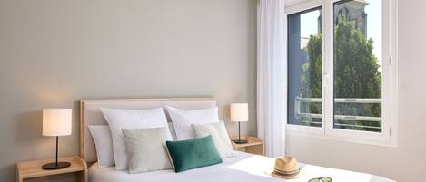 A neatly made bed with white linens and multiple pillows, a bedside table with a lamp, a window with a view of greenery and a building, and a hat and sunglasses on the bed.
