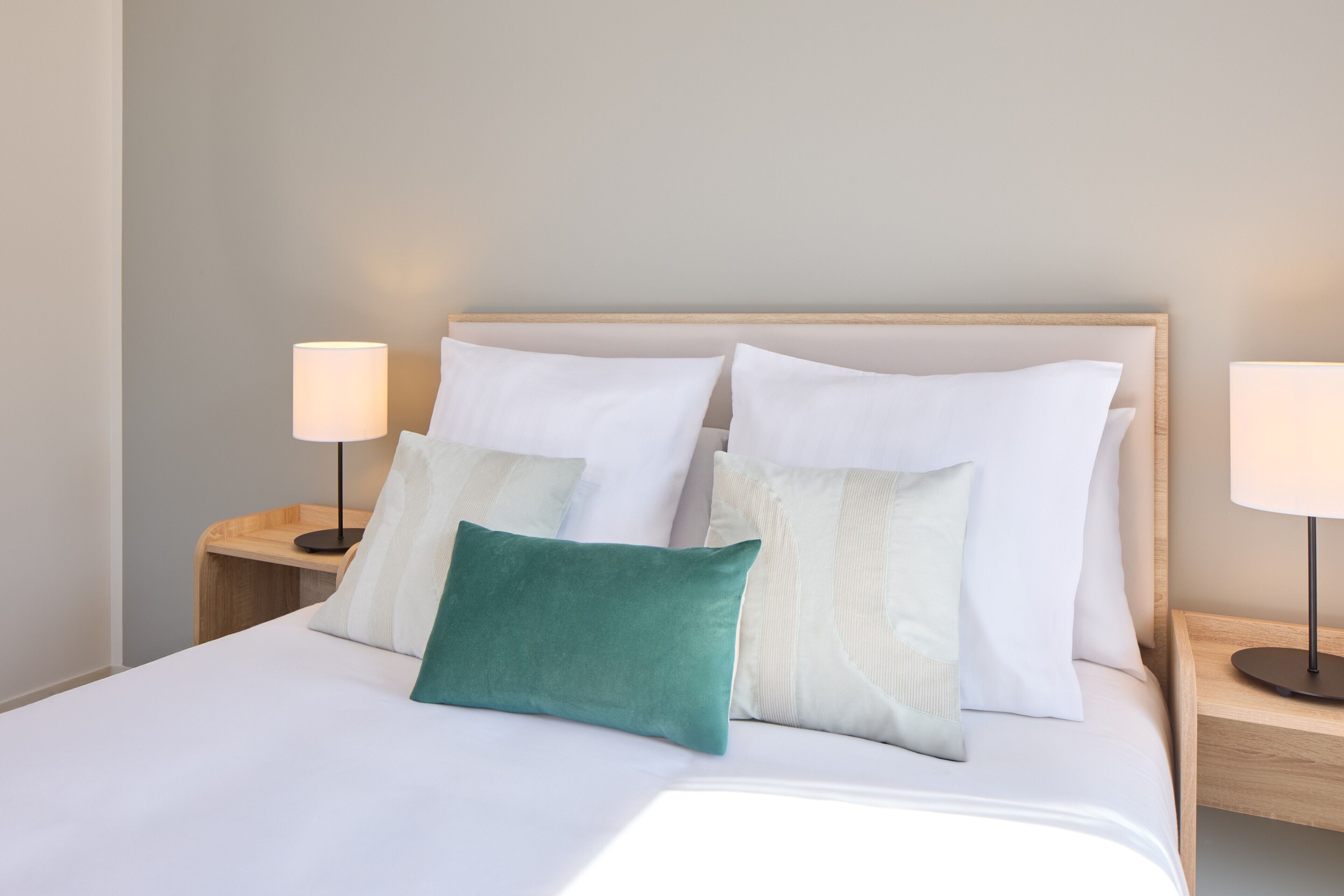 Una cama bien hecha con ropa de cama blanca y un almohadón verde azulado, flanqueada por dos mesitas de noche con lámparas, en una habitación con pared lisa y cabecera de madera.