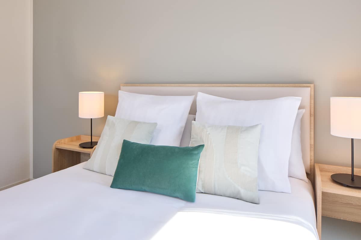 A neatly made bed with white bedding and a teal pillow, flanked by two bedside tables with lamps, in a room with a plain wall and a wooden headboard.