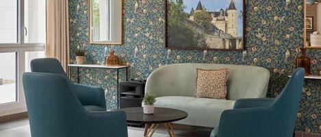 Una habitación con papel tapiz floral, un sofá, dos sillones, una mesa redonda, un espejo y un cuadro con la imagen de un edificio.