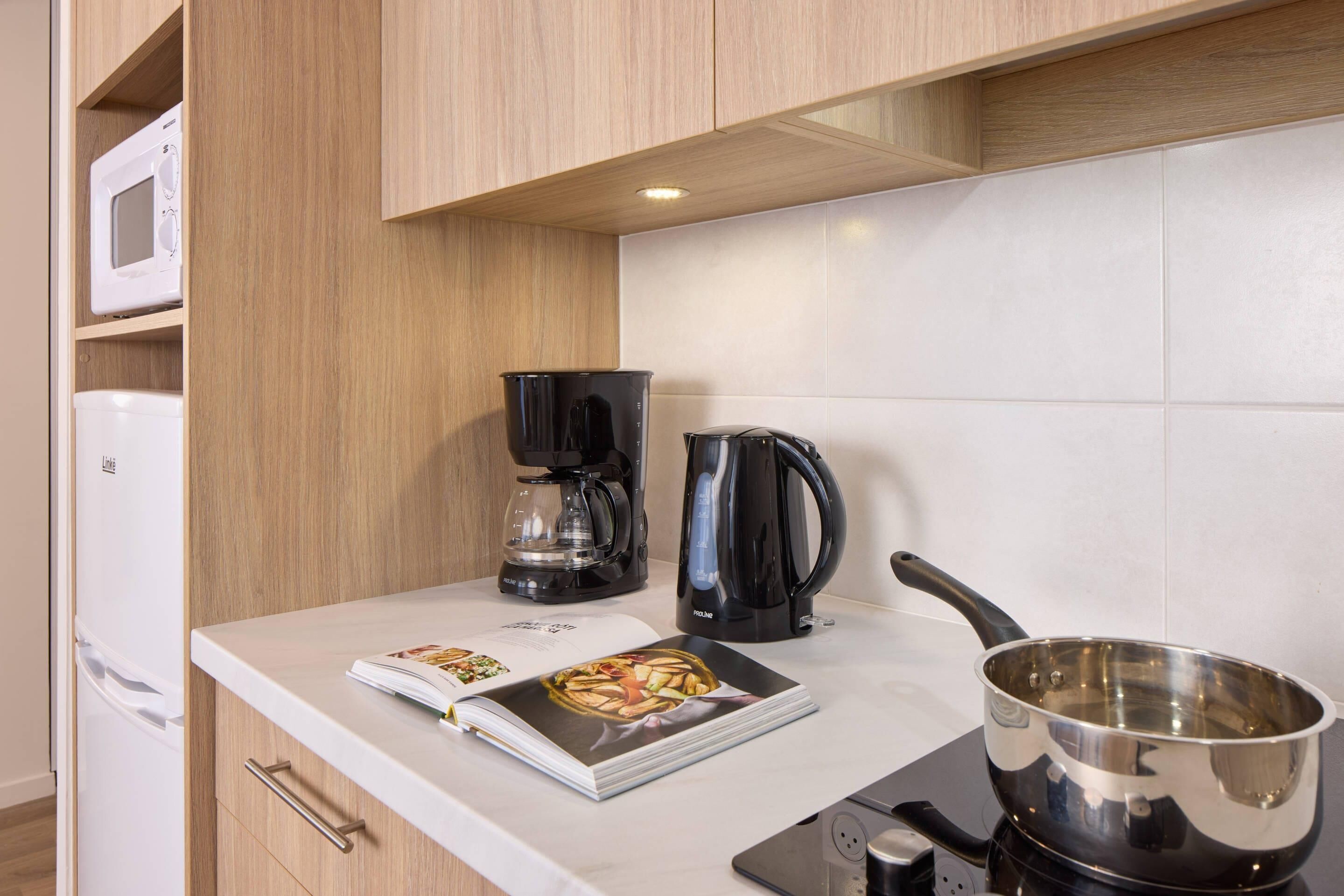 A kitchen with a microwave, coffee maker, kettle, and a cookbook on the counter.