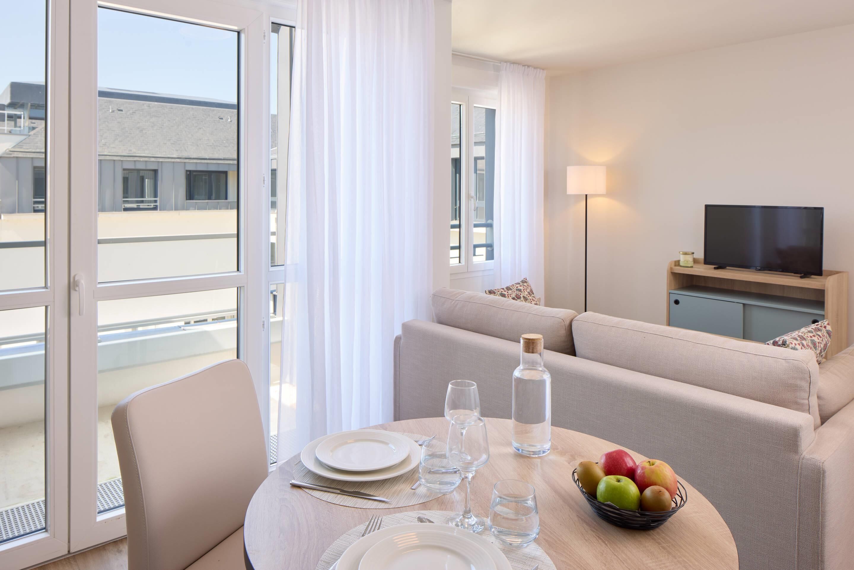 A modern hotel room with a dining area, a sofa, a television, and a view of the cityscape.
