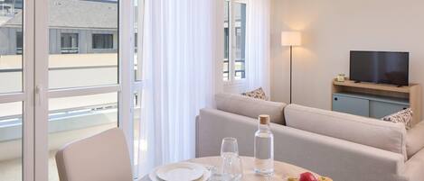 A modern hotel room with a dining area, a sofa, a television, and a view of the cityscape.