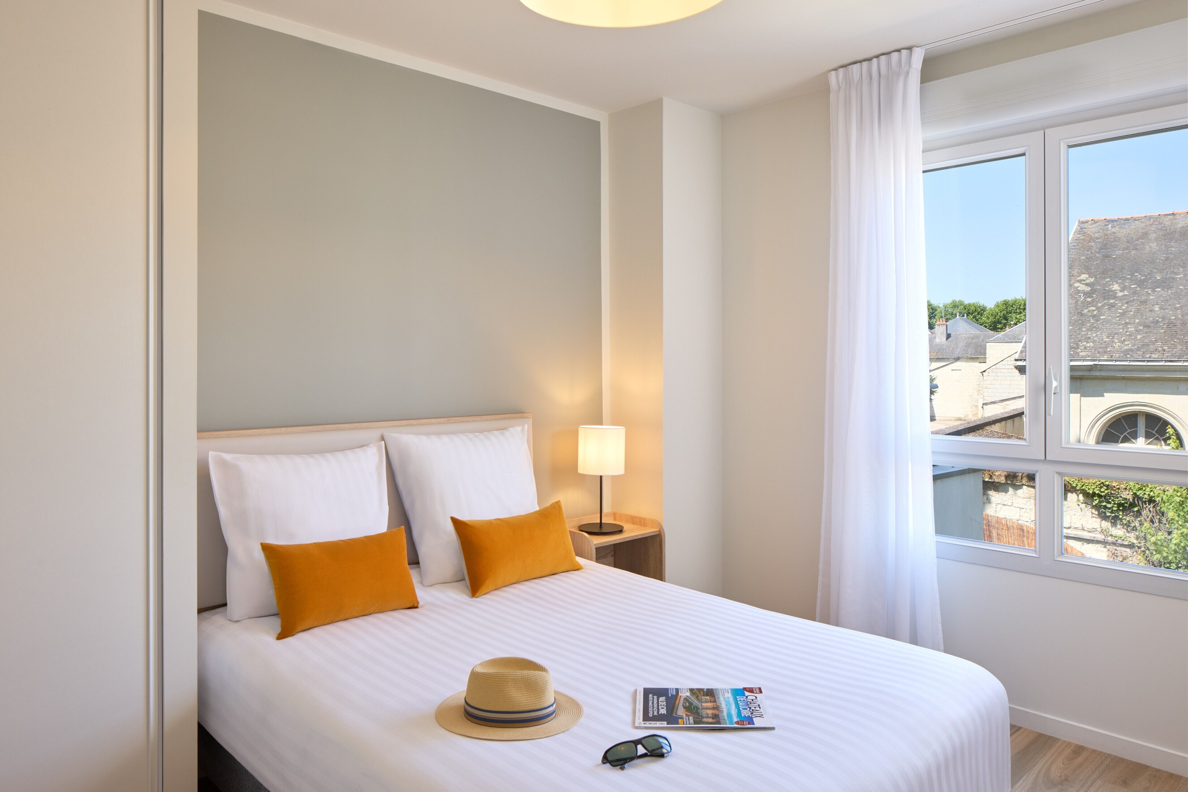 Habitación de hotel con cama, mesita de noche, lámpara, ventana con cortinas y vistas a edificios.