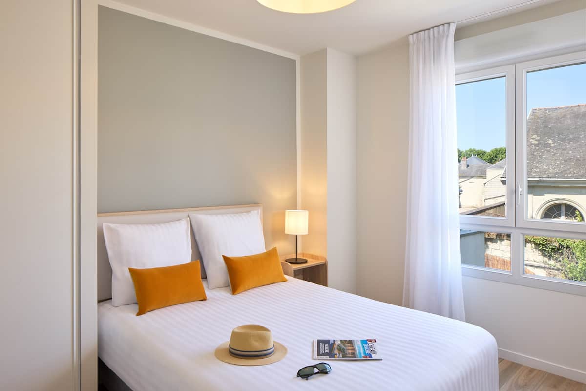 A hotel room with a bed, a nightstand, a lamp, a window with curtains, and a view of buildings outside.