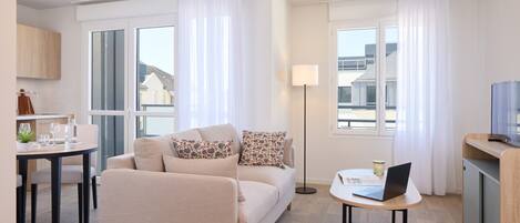 A modern living room with a sofa, a small round table with a laptop, and a TV on a wooden stand.
