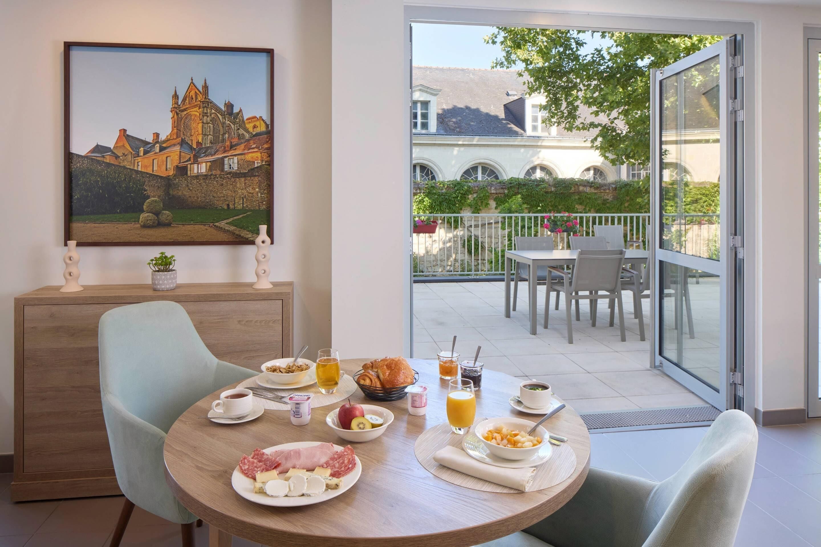 A dining area with a table set for breakfast, a painting of a castle on the wall, and a view of a courtyard with a building in the background.