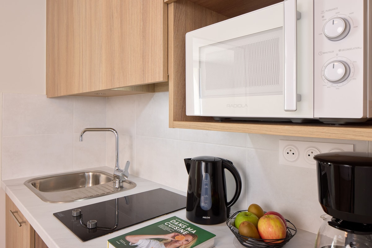 A kitchen with a microwave, kettle, sink, induction cooktop, and a bowl of fruit.