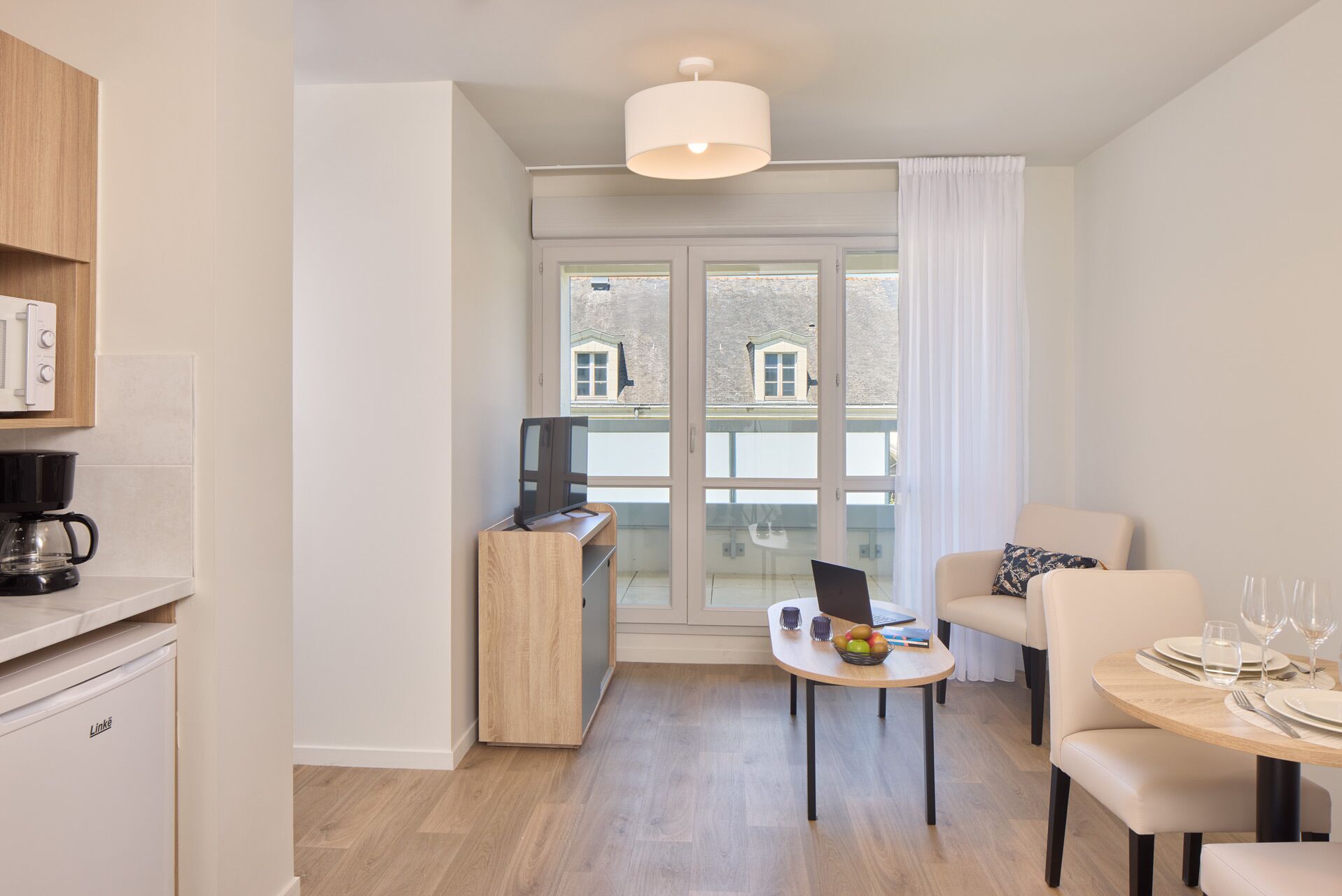 Una sala de estar moderna con comedor, una cocina equipada con cafetera y un ventanal con vistas a edificios.
