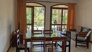A dining area with a glass-top table, wooden chairs, and a sofa. There is a refrigerator on the right and a window with curtains.