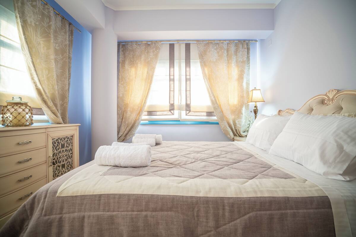 A neatly made bed with a quilted cover, a bedside table with a lamp, and curtains on a window.