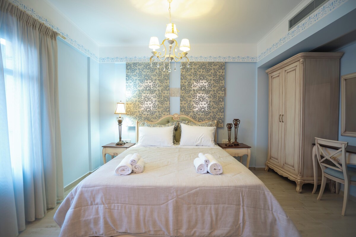 Un lit bien fait, agrémenté de serviettes pliées, un lustre et une grande armoire dans une pièce dont les murs sont recouverts d’un papier peint à motifs.