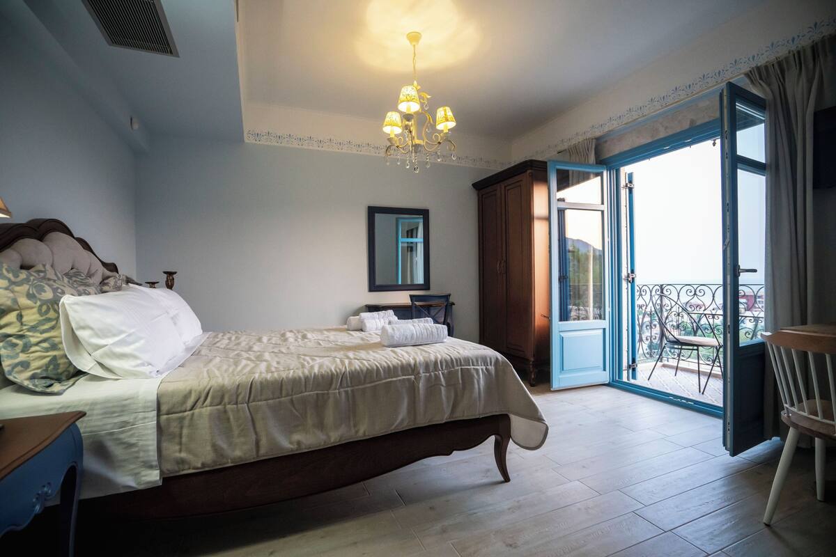 A bedroom with a bed, a chandelier, a mirror, and a balcony with a chair.