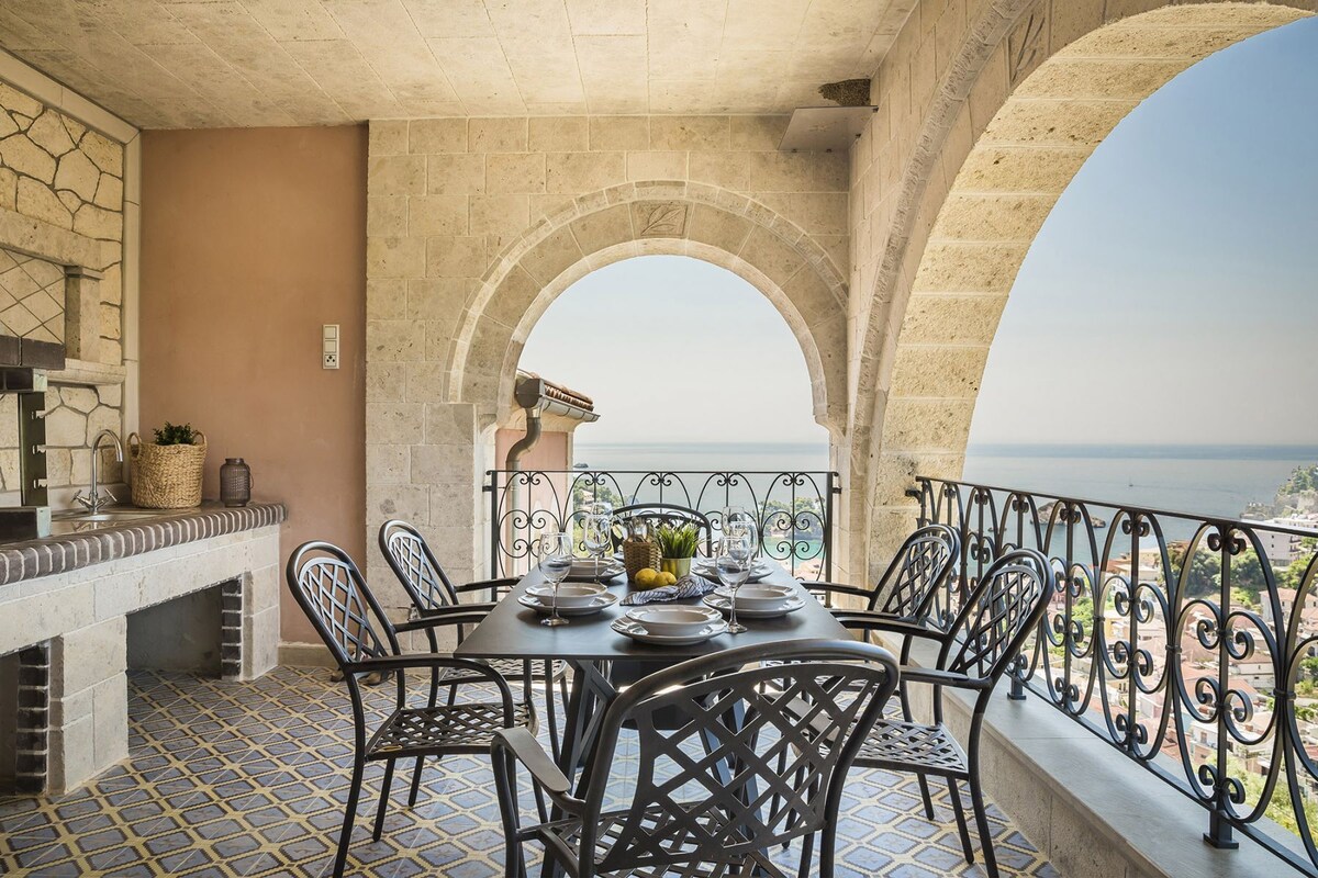 A balcony with a table set for dining, stone walls, and a view of the sea.