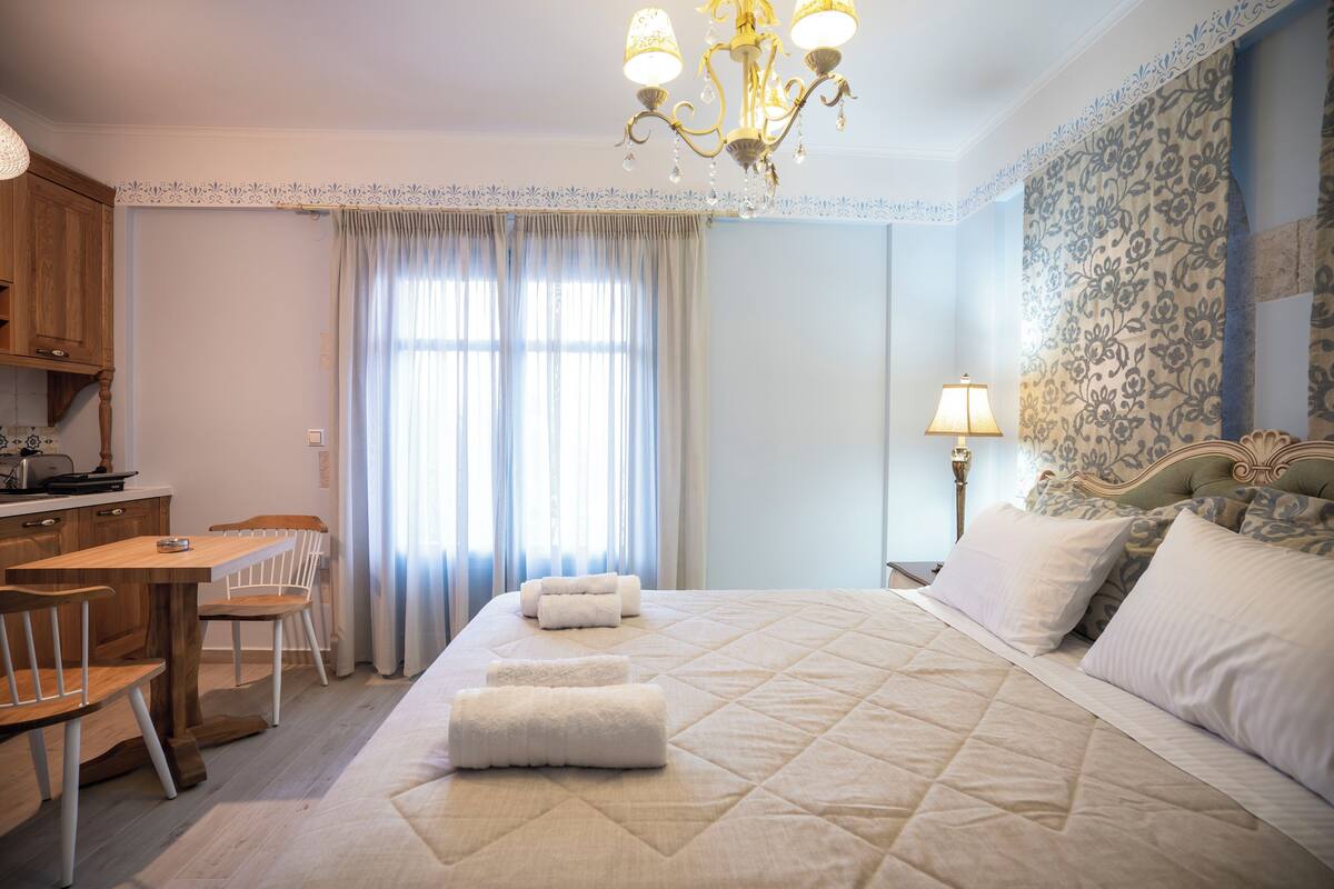 A hotel room with a bed, a desk, chairs, a chandelier, and a window with curtains.