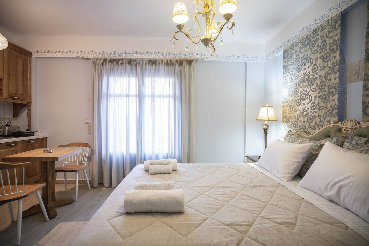 A bedroom with a bed, a desk, chairs, a chandelier, and a nightstand.