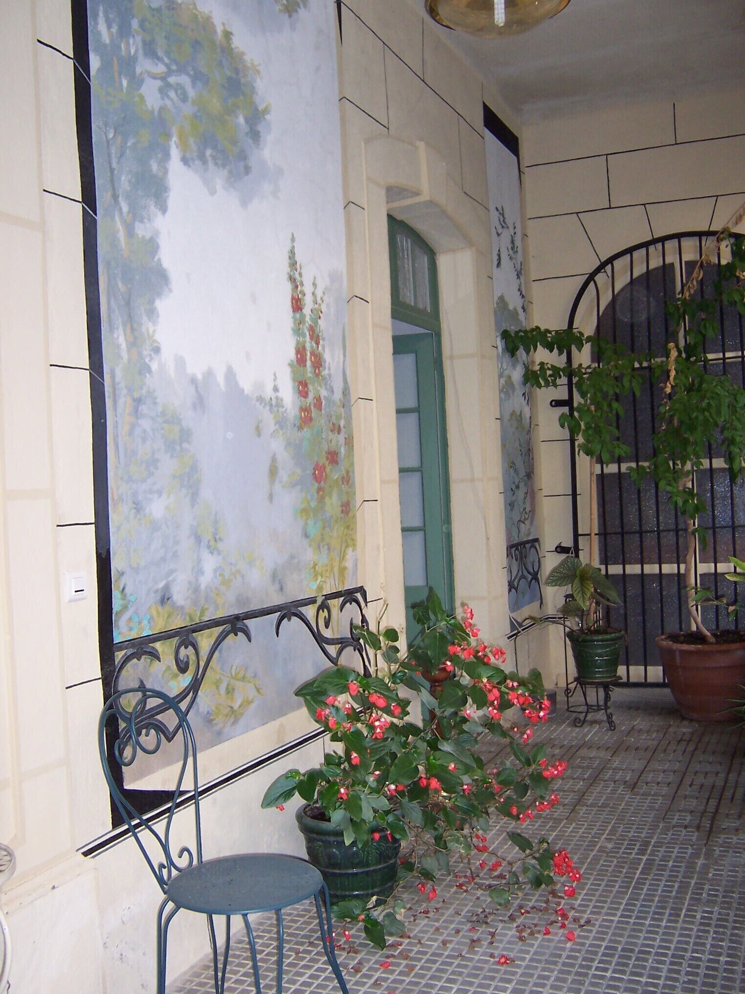Un balcone piastrellato con un murale, piante in vaso e una sedia in metallo.
