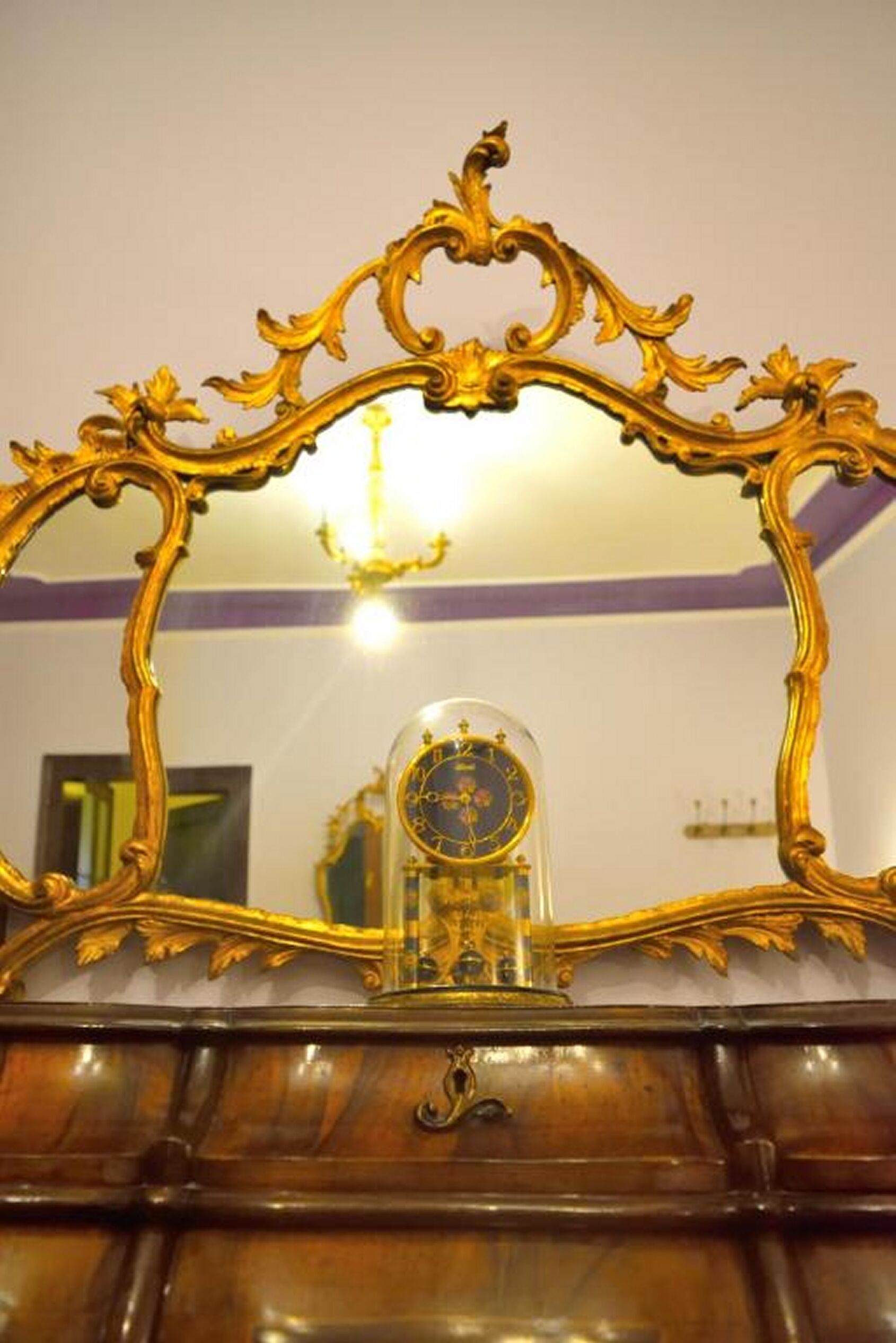 Ein Spiegel mit verziertem Goldrahmen, der einen Raum mit einer Kandelaber und einer Uhr reflektiert.